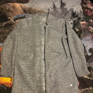 Grå kappa från Zink - Säljer en stilren grå kappa från Zink i storlek M. Den har en enkel design med långa ärmar och är perfekt för kyligare dagar. Kappan är gjord i ett mjukt material som ger en bekväm passform.