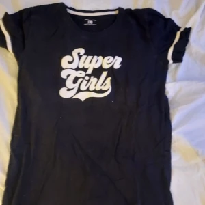 Svart t-shirt med texten 'Super Girls' - Snygg svart t-shirt med vit text 'Super Girls' på framsidan. T-shirten har korta ärmar med vita ränder vid ärmsluten. En supermysig T-shirt som passar både som pyjamas och vardags T-shirt!