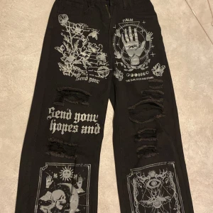 Svarta jeans med tryck och slitningar - Coola svarta jeans med unika tryck och slitningar. Byxorna har detaljerade grafiska mönster och text på framsidan, samt slitna partier för en edgy look. Perfekta för dig som vill sticka ut!