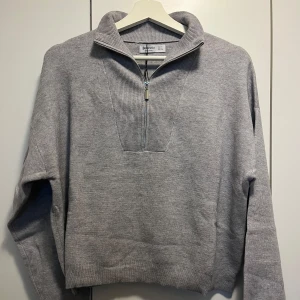 Grå stickad tröja från Stradivarius - Säljer en stilren grå stickad tröja från Zara med dragkedja vid halsen. Tröjan har en hög krage och långa ärmar, perfekt för kyligare dagar. Den är i en mjuk och bekväm kvalitet som passar till många olika outfits.