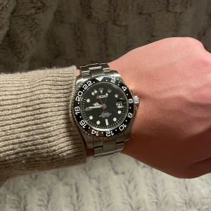 Seiko GMT Mod Svart - Svin snygg Seiko klocka med svart urtavla och datumvisning. 40mm.  Den har en silverfärgad boett och armband i rostfritt stål. Klockan har en tydlig 24-timmarsmarkering på den svarta ringen runt urtavlan. Klockan efterliknar den kända modellen GMT. Alla länkar tillkommer såklart vid köp. 