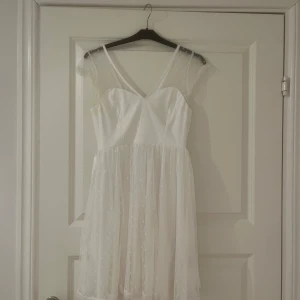 Vit klänning med spetsdetaljer -  Vit klänning med spetsdetaljer och prickigt mönster. Den har en v-ringad design med smala axelband och en knytrosett i ryggen. Perfekt för studenten!🌸