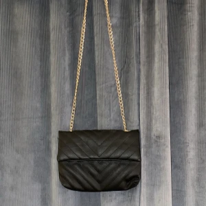 Svart axelväska med guld kedja - Svart axelväska med design och guldfärgad kedja som axelrem. Knapp som öppning till väskan.