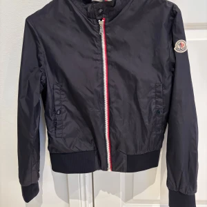 Mörkblå jacka från Moncler - Snygg mörkblå jacka från Moncler med dragkedja i rött och vitt. Jackan har långa ärmar och en Moncler-logga på ärmen. Perfekt för en stilren look.