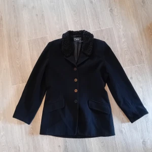 Svart ullkappa med pälsdetaljer - Säljer en svart kappa med snygga pälsdetaljer på kragen. Den har en klassisk design med knappar framtill och två fickor. Perfekt för hösten.Storlek 38 