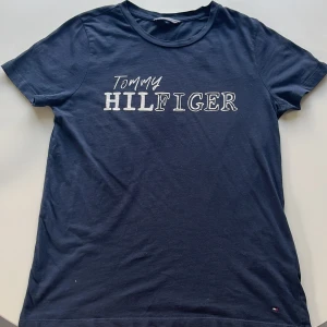 Mörkblå t-shirt från Tommy Hilfiger - Säljer en snygg mörkblå t-shirt från Tommy Hilfiger i storlek XS. Den har ett klassiskt tryck med märkesnamnet på framsidan och en liten logga nedtill. Perfekt för en casual look!
