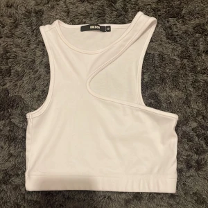 Vit asymmetrisk topp från Bik Bok - Säljer en supercool vit asymmetrisk topp från Bik Bok i storlek XS. Den är ärmlös och har en unik design med en snedskuren detalj över bröstet. Perfekt för sommarens alla fester eller bara en dag på stan. Materialet känns mjukt och skönt mot huden.