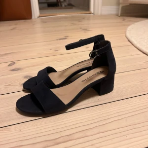 Svarta sandaler med klack från Tamaris - Snygga marinblåa klackar från Tamaris med en bekväm klack och justerbart spänne runt ankeln. Perfekta för både vardag och fest! De är gjorda i ett mjukt material som ser ut som mocka.