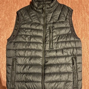 Svart dunväst med dragkedja - Säljer en svart dunväst som är perfekt för kyliga dagar. Västen har en dragkedja framtill och två praktiska fickor med dragkedjor. Den quiltade designen ger en snygg och modern look. Passar bra till både vardag och utomhusaktiviteter.