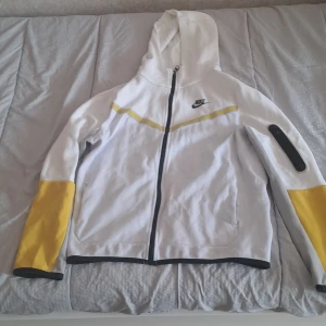 Vit och gul huvtröja från Nike - Säljer en vit och gul huvtröja från Nike i storlek XL. Tröjan har en dragkedja framtill och en huva. Den är vit med gula detaljer på ärmarna och en gul rand över bröstet. Det finns några fläckar på ena ärmen. Perfekt för en sportig look eller chill dagar.