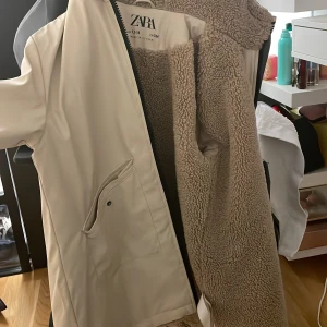 Beige jacka från Zara - Säljer en snygg beige jacka från Zara. Den har en mysig teddyfoder på insidan och en dragkedja framtill. Jackan har två stora fickor med knappar och en huva för extra värme. Perfekt för kyliga dagar!