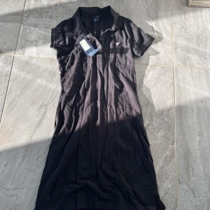 Svart klänning från GANT - Säljer en klassisk svart pikéklänning från GANT. Den har korta ärmar och en stilren krage med knappar framtill. Klänningen är i nyskick och passar perfekt för både vardag och festliga tillfällen.