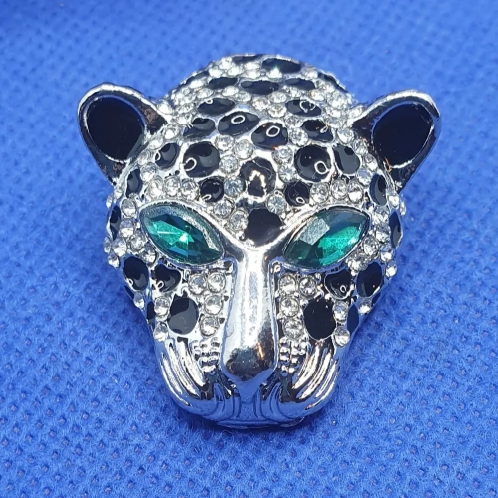 Den här broschen i form av ett leopardhuvud med gröna ögon har silverfärgad metall och små transparenta kristaller, mått är ca 3.7 × 3.5 cm. Denna fungerar även att använda som hänge till halsband då den har en ögla på baksidan. . Accessoarer.