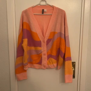 Färgglad kofta från H&M Divided - Säljer en superfin och färgglad kofta från H&M Divided i storlek L. Den har ett coolt mönster i rosa, orange och lila nyanser. Perfekt för att liva upp garderoben! Koftan är stickad och har långa ärmar med knappar framtill. Passar perfekt för både vår och höst.