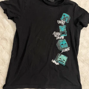 Svart Minecraft t-shirt - Säljer en svart t-shirt med coolt Minecraft-tryck från Kappahl i storlek 158/164. Den har korta ärmar och är perfekt för alla fans av spelet. Trycket visar olika karaktärer som Creeper och Zombie i blått och grönt. Perfekt för vardagsbruk eller gamingkvällar! 🎮