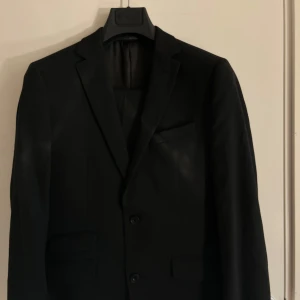 Svart kavaj från Riley - Säljer en stilren svart kavaj från Riley i storlek Slim Fit. Perfekt för både formella tillfällen och fester. Kavajen har en klassisk design.