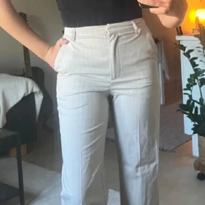 Beige randiga kostymbyxor från BikBok  - Snygga beige kostymbyxor från BikBok  i storlek 34/XS. De har ett diskret randigt mönster och är högmidjade för en elegant look. Perfekta för både kontoret och festliga tillfällen. Byxorna har en rak passform och stängs med dragkedja och knapp framtill.