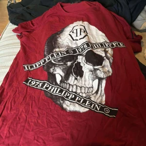 Röd t-shirt med dödskalle från Philipp Plein - Säljer en cool röd t-shirt från Philipp Plein med en stor dödskalletryck på framsidan. T-shirten har korta ärmar och är perfekt för en avslappnad stil. Den är i bra skick.