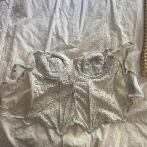 Vit spetskorsett - Säljer en elegant vit korsett med vacker spetsdetalj. Den har justerbara axelband och en knäppning fram. Perfekt för speciella tillfällen eller som en snygg detalj under kläder. Passar bra till både fest och vardag.