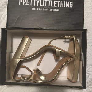 Guldiga klackskor från PrettyLittleThing - Snygga guldiga klackskor från PrettyLittleThing med en bred klack och justerbart spänne runt ankeln. Perfekta för festliga tillfällen! Skorna är i syntetmaterial och har en öppen tådesign. De är i välanvänt skick med lite slitage på sulorna. Storlek 38