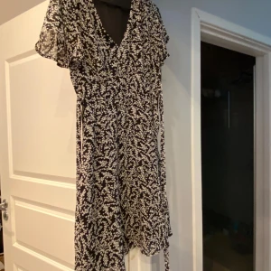 Svart och vit blommig klänning - Säljer en superfin svart klänning med vita blommor. Den har en v-ringad hals och korta ärmar med volangdetaljer. Perfekt för både vår och sommar! Klänningen är i nyskick och passar perfekt till både vardag och fest.