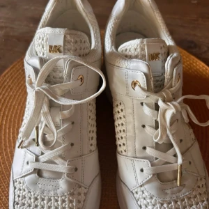 Vita sneakers från Michael Kors - Säljer ett par snygga vita sneakers från Michael Kors. De har en cool flätad design på sidorna och är gjorda i skinn. Skorna har snörning och guldiga detaljer som ger en lyxig touch. Perfekta för både vardag och fest!