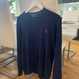 Mörkblå tröja från Polo Ralph Lauren - Säljer en snygg mörkblå tröja från Polo Ralph Lauren i storlek M. Den har en klassisk design med det ikoniska logotypbroderiet i orange på bröstet. Perfekt för både vardag och lite finare tillfällen. Tröjan är i bra skick lite uttöjd runt magen men inget som påverkar utseendet och superbekväm att ha på sig