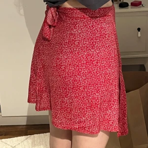 Röd omlottkjol med knytband - Säljer en superfin röd omlottkjol med små vita blommor. Kjolen har ett knytband i midjan som ger en söt detalj och perfekt passform. Den är kort och passar perfekt till sommaren eller en festlig kväll. 
