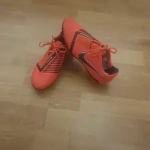 Säljer ett par coola orangea Nike fotbollsskor i bra skick. De har snörning och en snygg svart detalj på sidan. Perfekta för fotbollsträningen och ger bra grepp på planen. Skorna är bekväma och lätta, vilket gör dem idealiska för snabba rörelser.