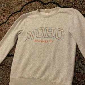 Grå sweatshirt med NOHO-tryck - Säljer en snygg grå sweatshirt med texten 'NOHO New York City' tryckt på framsidan. Tröjan har en klassisk rund hals och långa ärmar, perfekt för en avslappnad stil. Den är i bra skick och passar perfekt för både höst och vår. Perfekt för dig som vill ha en stilren och bekväm tröja!