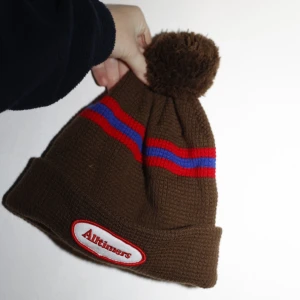 Brun mössa med tofs från Alltimers - Säljer en snygg brun mössa från Alltimers med en stor tofs på toppen. 