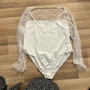Vit body med transparenta ärmar - Säljer en superfin vit body med transparenta, prickiga ärmar. Den har en elegant och stilren design med långa ärmar och volangdetaljer vid ärmsluten. Perfekt för en festlig kväll eller en stilfull vardag. Passar bra till både kjol och byxor!