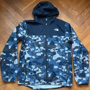 Säljer en snygg och praktisk vindjacka från The North Face i blått och svart camouflagemönster. Jackan har en dragkedja framtill och en justerbar huva. Storlek XL kids motsvarar XS/S. Upp till 175 cm ungefär. Perfekt för höst och vår när vädret är blåsigt. Den är i mycket bra skick, inga hål eller fläckar! 🌟