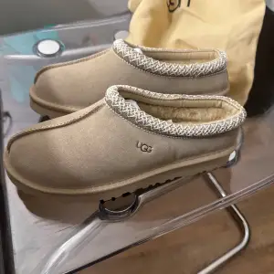 Säljer ett par supermysiga beige tofflor från UGG. De har en mjuk insida och en snygg stickad kantdetalj runt öppningen. Perfekta för att hålla fötterna varma under kalla dagar. De är i mycket bra skick och kommer med en praktisk förvaringspåse.