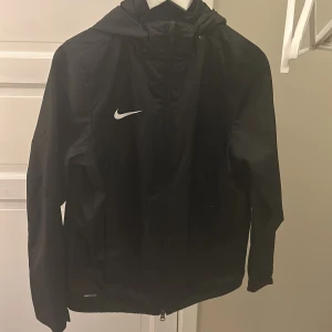 Svart vindjacka från Nike - Säljer en svart vindjacka från Nike i storlek XL. Jackan har en huva och dragkedja framtill, perfekt för blåsiga dagar. Den är i mycket bra skick och har Nike Shield-teknologi för extra skydd mot väder och vind.
