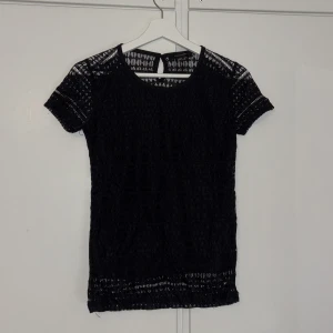 Svart spetsblus - Snygg svart spetsblus med korta ärmar och ett elegant mönster. Perfekt för både vardag och fest. Blusen har en fin detalj med knäppning i nacken och är i mycket bra skick.