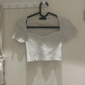 Vit ribbad croppad topp från Zara - Säljer en superfin vit ribbad topp från Zara. Den är croppad och har korta ärmar, perfekt för sommardagar. Toppen har en söt, hjärtformad urringning som ger en feminin touch. Passar både till vardags och fest!