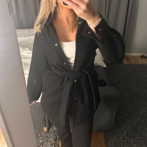 Svart kappa  - Snygg svart kappa med snörning och knappar framtill. Perfekt för höst och vår. Långärmad design som ger en stilren look.