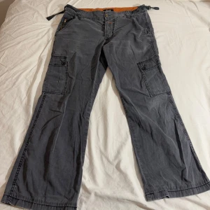 Vintage Grå cargobyxor från Dolce & Gabbana - Snygga vintage grå cargobyxor från Dolce & Gabbana med flera praktiska fickor och knytband i midjan. Byxorna har en avslappnad passform och är perfekta för en cool och bekväm stil. Märkeslogga synlig både på insidan och baksidan. Materialet är 100% cotton. 