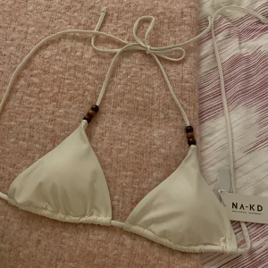 Vit bikiniöverdel från NA-KD - Snygg vit bikiniöverdel från NA-KD med triangelformade kupor och dekorativa träpärlor på banden. Perfekt för en stilren strandlook. Helt oanvänd, lapp kvar