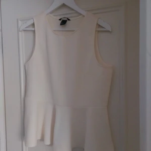 Vit ärmlös topp med volang - Säljer en stilren vit ärmlös topp med volangdetalj i nederkant. Perfekt för en elegant och avslappnad look. Toppen har en rund halsringning och är tillverkad i ett lätt och bekvämt material.