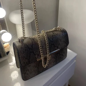 Ormmönstrad axelväska med kedja - Snygg axelväska med ormmönster och guldfärgad kedja. Väskan har en elegant låsdetalj framtill och en stilren design som passar perfekt för en trendig look. Perfekt för att ge din outfit en extra touch av stil.