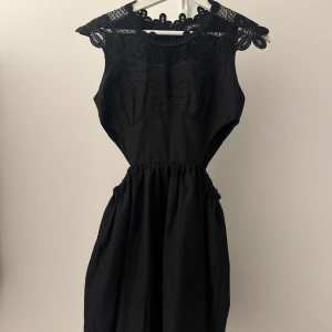 Svart klänning med spetsdetaljer - Elegant svart klänning med vackra spetsdetaljer vid axlarna och halsen. Klänningen har en smickrande passform med en markerad öppen midja och är ärmlös. Perfekt för en stilren look.