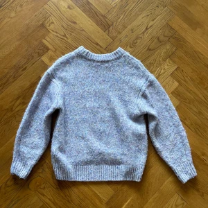 Ljuslila stickad tröja - Mysig ljuslila stickad tröja med en melerad effekt i olika pastellfärger. Tröjan har långa ärmar och ribbstickade muddar vid ärmslut och nederkant. 