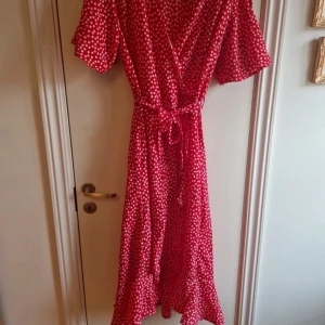 Röd prickig omlottklänning - Charmig röd omlottklänning med vita prickar och knytband i midjan. Klänningen har korta ärmar och en v-ringad design som ger en feminin touch. Perfekt för sommarens varma dagar.