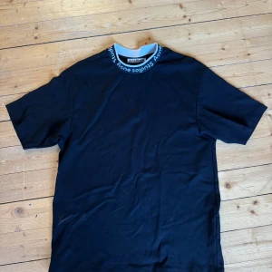 Svart t-shirt från Acne Studios - Snygg svart t-shirt från Acne Studios med logotyp runt halsringningen. Perfekt för en stilren look. Tillverkad i mjukt material för extra komfort. Stor i storleken