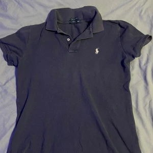 Svart pikétröja från Ralph Lauren (THE SKINNY V) - Säljer en klassisk svart/mörkblå pikétröja från Ralph Lauren i storlek S (The skinny version st L). Tröjan är tillverkad i 100% bomull och har en broderad logotyp på bröstet. Perfekt för en stilren look.