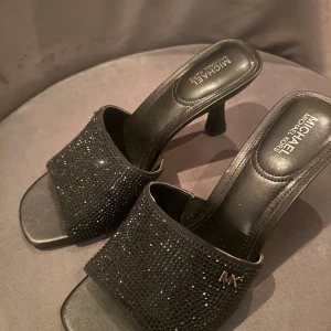 Svarta klackskor från Michael Kors - Eleganta svarta klackskor från Michael Kors med glittrande detaljer och öppen tå. Skorna har en stilren design med en liten klack och är perfekta för att ge en touch av glamour till din outfit.