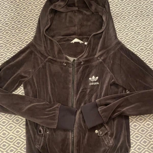Grå-svart velourjacka från Adidas - Adidas zip up som är i bra skick i storlek 38/40. Har knytning bak på luvan. KÖP ENDAST VIA POSTNORD 