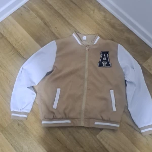 Beige och vit bomberjacka med bokstavsdetalj - Snygg bomberjacka i beige och vitt med en stor bokstav 'A' på bröstet. Jackan har vita ärmar och ribbade muddar i beige och vitt. Den stängs med dragkedja och har två fickor framtill.